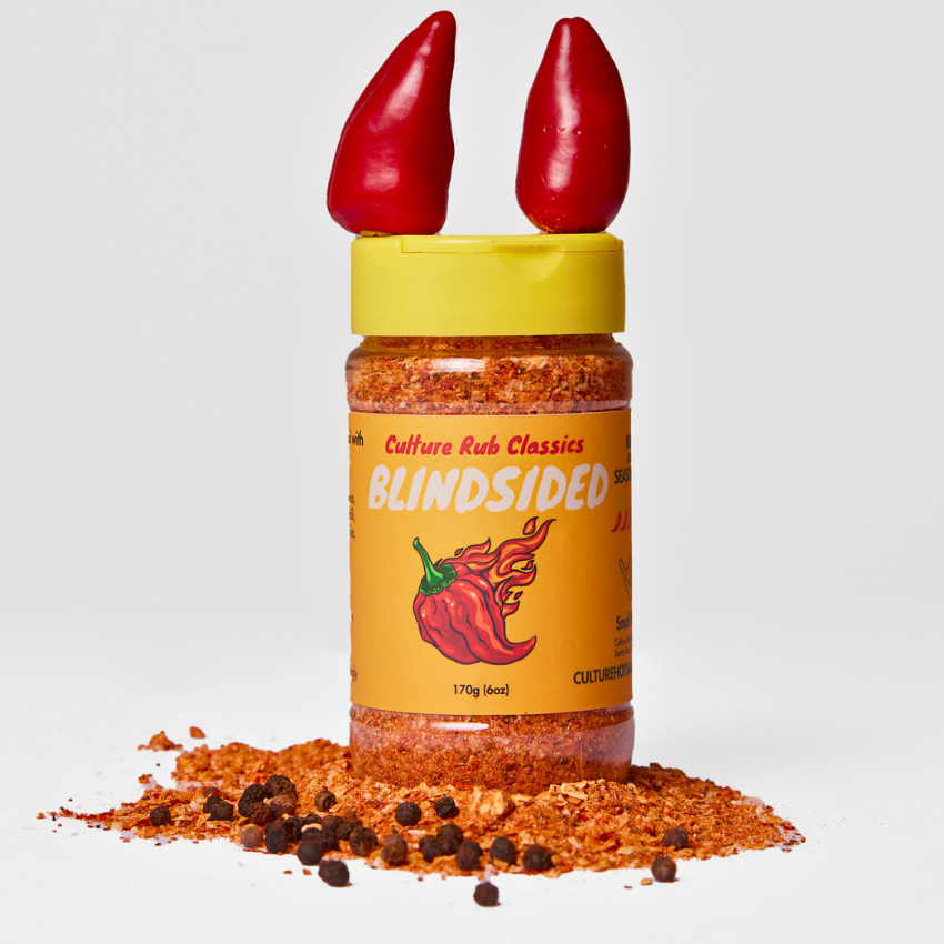 A bottle of Culture Rub Classics Blindsided hot sauce rub stands on a pile of red seasoning and peppercorns against a light gray background. The bottle has a yellow cap with two red chili peppers sticking out of it, and the label is orange with a red chili pepper graphic.