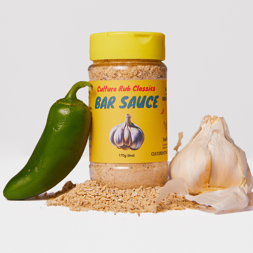 A bottle of Culture Rub Classics Bar Sauce stands on a pile of coarse seasoning, flanked by a green jalapeño pepper and a whole garlic bulb on a clean white background.
