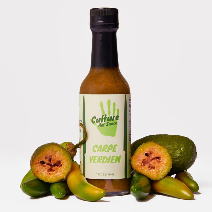 A bottle of Culture Hot Sauce Carpe Verdiem hot sauce centered on a white background, surrounded by fresh green chilies and sliced guavas.