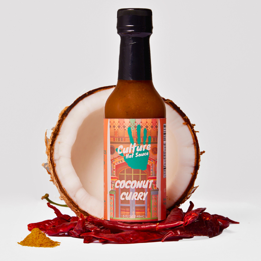 A bottle of Culture Hot Sauce, Coconut Curry flavor, styled with a halved coconut, scattered red chili peppers, and a small pile of yellow curry powder on a light gray background.