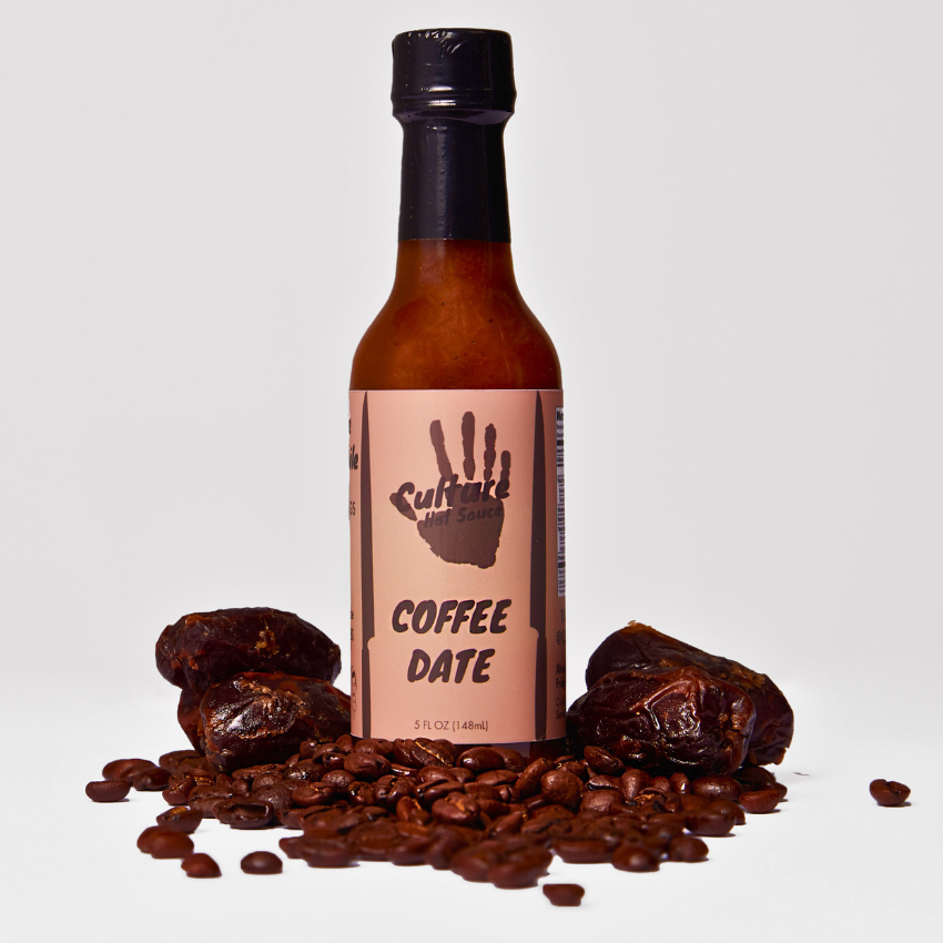 A bottle of Cultivate Coffee Date hot sauce, featuring a light brown label with a handprint logo, surrounded by coffee beans and dates on a light gray background.