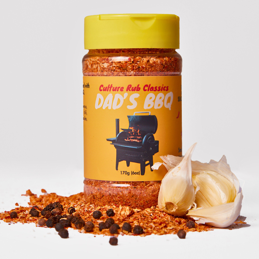 A bottle of Culture Rub Dad's BBQ seasoning with a yellow cap and yellow label, styled with scattered seasoning, peppercorns, and a garlic bulb on a white background.