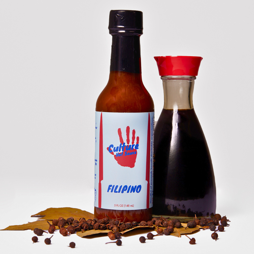 A bottle of Culture Hot Sauce Filipino hot sauce stands next to a glass carafe of dark soy sauce, both set on a light gray background with dried bay leaves, peppercorns, and garlic cloves in front. Based on adobo.