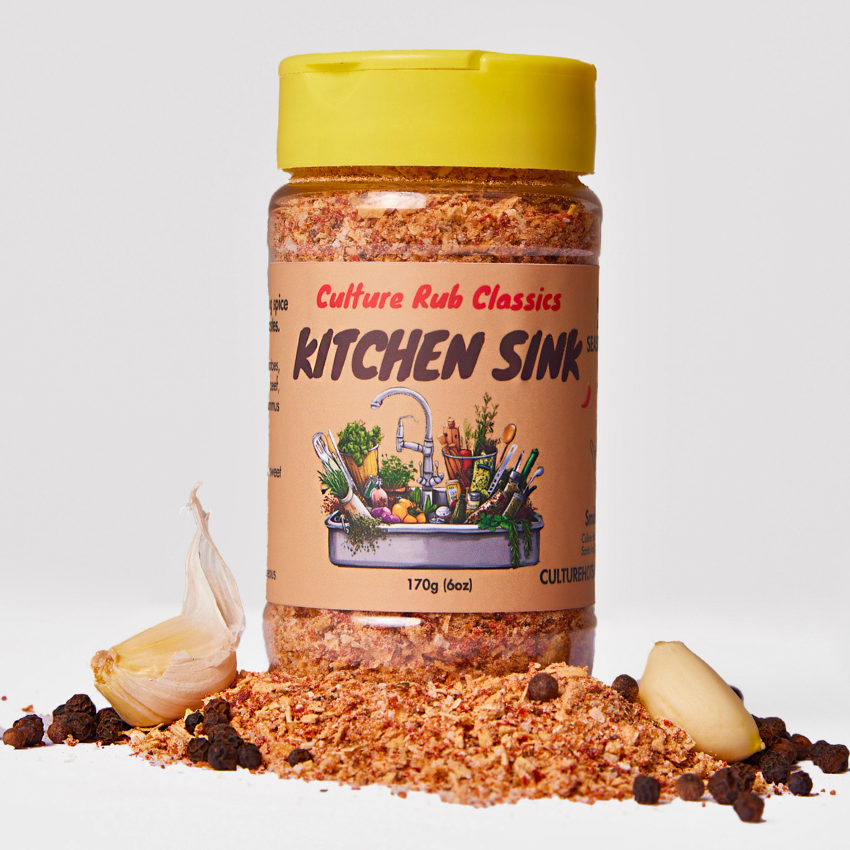 A bottle of Culture Rub Classics Kitchen Sink seasoning with a bright yellow cap, surrounded by loose seasoning, garlic cloves, and peppercorns on a clean white background.