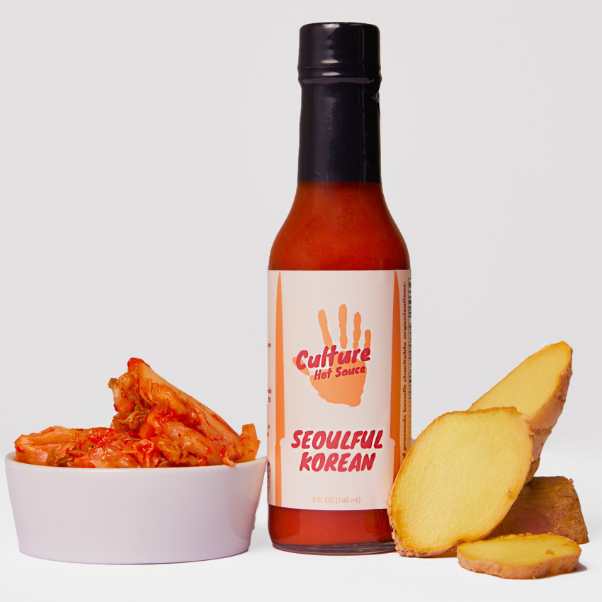 A bottle of Culture Hot Sauce Seoulful Korean hot sauce stands next to a small white bowl of kimchi and slices of ginger on a clean white background.