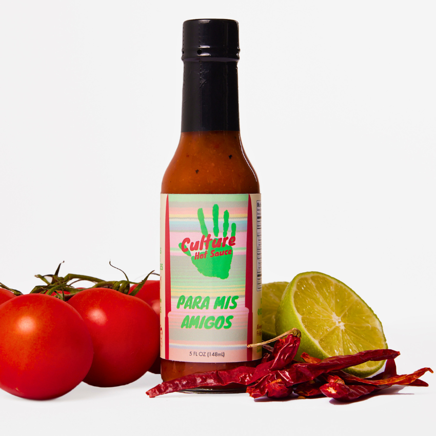 A bottle of Culture Hot Sauce labeled 'Para Mis Amigos' stands centered on a white background, surrounded by fresh ingredients including red tomatoes, a lime half, red chili peppers, and a green handprint on the label.