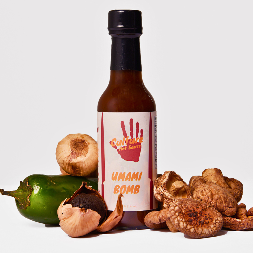 A bottle of Umami Bomb hot sauce centered on a light gray background, surrounded by ingredients including a jalapeño, garlic, and mushrooms. The bottle has a black cap and a white label with a red handprint and orange text.